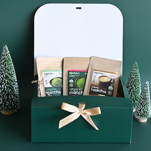 May include: A green gift box with a white lid, tied with a gold bow, contains three packages of Aprka organic matcha and hojicha tea. Two small decorative Christmas trees are on the right side of the box.