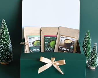 May include: A green gift box with a white lid, tied with a gold bow, contains three packages of Aprka organic matcha and hojicha tea. Two small decorative Christmas trees are on the right side of the box.