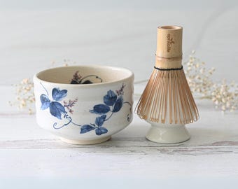 Puede incluir: Un cuenco de matcha de cerámica con diseños de hojas azules y un batidor de bambú. El cuenco es de color blanco roto con hojas azules y detalles rosas. El batidor es de bambú y tiene una base blanca.