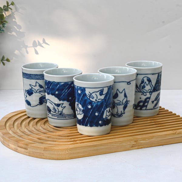 May include: Set of six white ceramic teacups with blue and white cat illustrations. The cups are arranged on a bamboo tray.