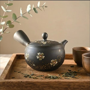 May include: A black ceramic teapot with a side handle and a lid, decorated with white and red floral designs. The teapot sits on a wooden tray with two matching cups and loose tea leaves.