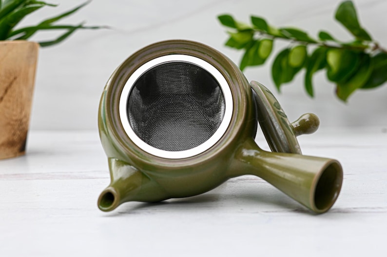 Puede incluir: Una tetera de cer&aacute;mica verde con un colador de acero inoxidable. La tetera tiene una tapa y un pico. La tetera est&aacute; sobre una superficie blanca.