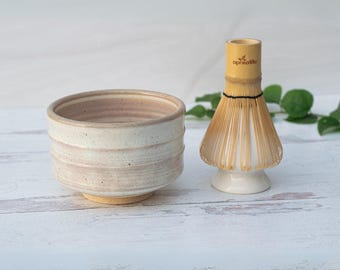 May include: A white ceramic matcha bowl with a light pink glaze and a bamboo matcha whisk with a white ceramic base. The whisk has a black string tied around the bamboo handle and the text "aprikalife" printed on the handle.