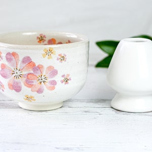 May include: A white ceramic teacup with a floral pattern in shades of pink and yellow. The cup is sitting on a white wooden surface next to a white ceramic tea whisk holder.