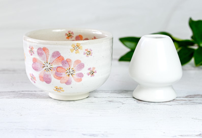 May include: A white ceramic teacup with a floral pattern in shades of pink and yellow. The cup is sitting on a white wooden surface next to a white ceramic tea whisk holder.