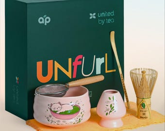 Op de afbeelding: Een roze matcha-kom met een kattenillustratie, een bamboe garde, een roze theekopje, een bamboe lepel en een zeef. De set is verpakt in een groene doos met de tekst "Unfurl" en "United by Tea".
