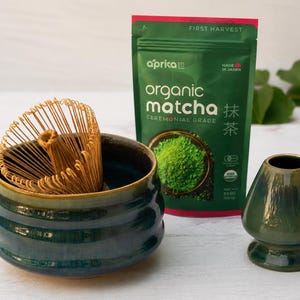 May include: A matcha tea set featuring a green ceramic bowl with a bamboo whisk, a green ceramic tea bowl, and a package of organic matcha powder. The matcha powder package is green with the words "organic matcha" and "ceremonial grade".
