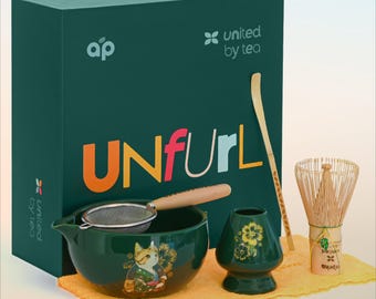 Puede incluir: Una caja verde con el texto "UNFURL" en letras de colores y el logotipo "united by tea". La caja contiene un cuenco de té de cerámica verde con un diseño de gato, un batidor de bambú, una cuchara de té de bambú, una taza de té de cerámica verde con diseños florales y un colador verde con un mango de madera.