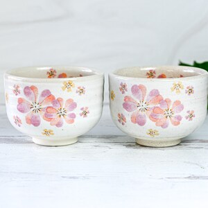 May include: Two white ceramic bowls with pink and yellow floral designs. The bowls are on a white wooden surface.