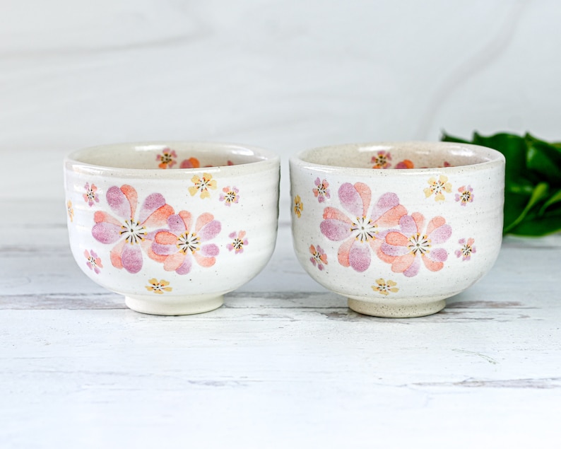 May include: Two white ceramic bowls with pink and yellow floral designs. The bowls are on a white wooden surface.