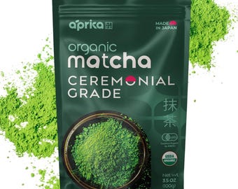 May include: A dark green bag of organic matcha powder with the brand name "aprika" and the text "CEREMONIAL GRADE." The bag also states "MADE IN JAPAN" and "USDA ORGANIC." The net weight is 3.5 OZ (100g). Green matcha powder is scattered around the bag.