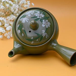 Puede incluir: Una tetera de cer&aacute;mica verde con un dise&ntilde;o floral blanco. La tetera tiene una tapa con un peque&ntilde;o agujero en la parte superior y un pico en el lateral.