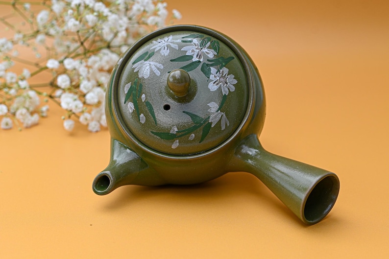 Puede incluir: Una tetera de cer&aacute;mica verde con un dise&ntilde;o floral blanco. La tetera tiene una tapa con un peque&ntilde;o agujero en la parte superior y un pico en el lateral.