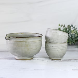 May include: A set of three ceramic bowls in a light gray color with a speckled glaze. The bowls are stacked on top of each other, with the largest bowl at the bottom and the smallest bowl at the top. The bowls have a slightly rounded shape and a smooth, matte finish.