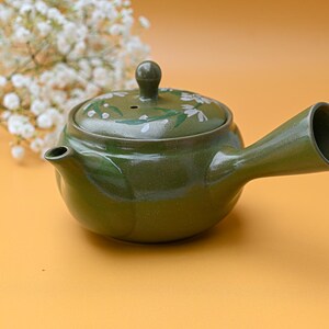 Puede incluir: Una tetera de cer&aacute;mica verde con un dise&ntilde;o floral blanco. La tetera tiene un pico y una asa. La tetera est&aacute; sobre una superficie amarilla.