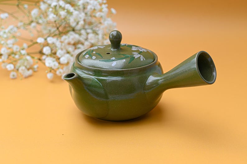 Puede incluir: Una tetera de cer&aacute;mica verde con un dise&ntilde;o floral blanco. La tetera tiene un pico y una asa. La tetera est&aacute; sobre una superficie amarilla.