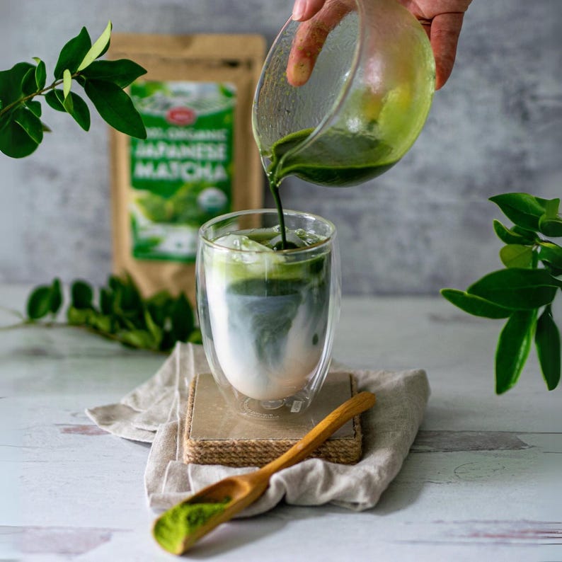 Puede incluir: Un vaso de doble pared con hielo y un l&iacute;quido lechoso se est&aacute; llenando con t&eacute; matcha verde. Una cuchara de madera con polvo de matcha est&aacute; sobre un pa&ntilde;o de lino. Una bolsa de Matcha japon&eacute;s 100% org&aacute;nico est&aacute; en el fondo.