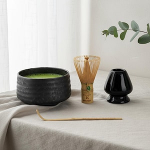 Può includere: Un set da tè matcha con una ciotola in ceramica nera piena di tè verde, un frullino di bambù, un portafrullino in ceramica nera e un cucchiaio di bambù. Il set è disposto su una tovaglia di lino color neutro.