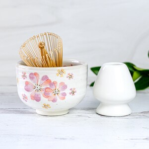 May include: A white ceramic tea bowl with a pink floral design and a bamboo whisk inside. A white ceramic tea scoop is next to the bowl.