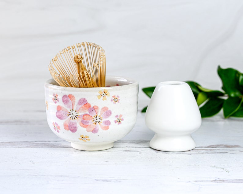 May include: A white ceramic tea bowl with a pink floral design and a bamboo whisk inside. A white ceramic tea scoop is next to the bowl.