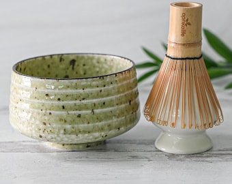 May include: A green ceramic matcha bowl with a speckled glaze and a bamboo matcha whisk with a white ceramic base. The whisk has the text "aprikaliife" printed on the handle.