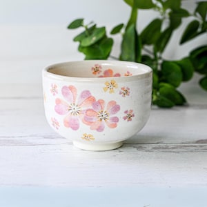 May include: A white ceramic teacup with a floral design. The cup has pink and yellow flowers on a white background.