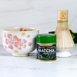 May include: A white ceramic bowl with a floral design, a green can of organic Japanese matcha powder, and a bamboo whisk with a white ceramic base.