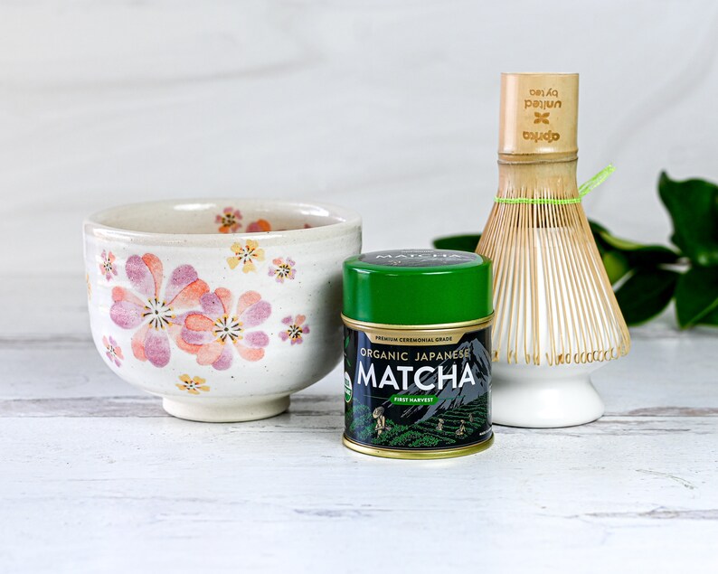 May include: A white ceramic bowl with a floral design, a green can of organic Japanese matcha powder, and a bamboo whisk with a white ceramic base.