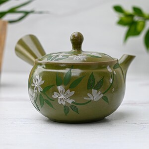 Puede incluir: Una tetera de cer&aacute;mica verde con un dise&ntilde;o floral blanco. La tetera tiene una tapa y un pico. La tetera est&aacute; sobre una superficie blanca.
