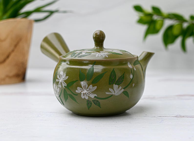 Puede incluir: Una tetera de cer&aacute;mica verde con un dise&ntilde;o floral blanco. La tetera tiene una tapa y un pico. La tetera est&aacute; sobre una superficie blanca.