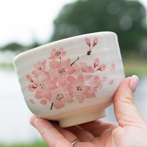 Puede incluir: Cuenco de cerámica color crema con diseño de flores de cerezo rosas. El cuenco tiene una forma ligeramente acampanada y un pequeño pie. Las flores están pintadas en el lateral del cuenco.