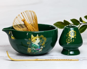 May include: A green ceramic matcha bowl with a gold cat design, a bamboo whisk, a matching green ceramic matcha scoop, and a bamboo matcha whisk.
