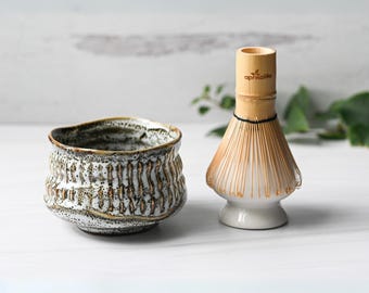 May include: A ceramic matcha bowl with a textured, wavy design in shades of white, brown, and gray sits next to a bamboo matcha whisk. The whisk has a white base and a light brown whisk head. The brand name "apricalife" is printed on the whisk.