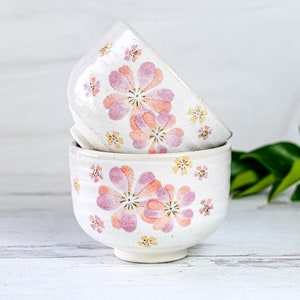 May include: Two white ceramic bowls with a floral pattern. The bowls are stacked on top of each other. The pattern features pink and yellow flowers on a white background.