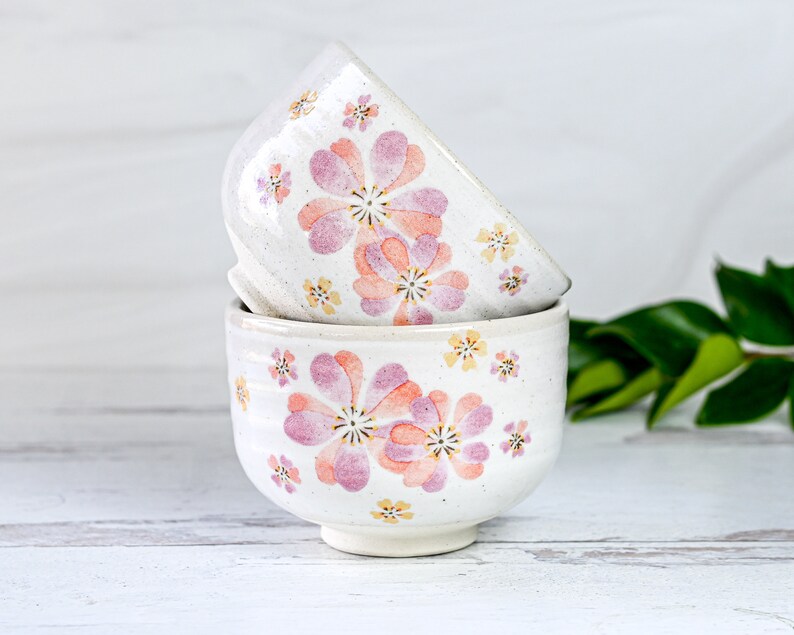 May include: Two white ceramic bowls with a floral pattern. The bowls are stacked on top of each other. The pattern features pink and yellow flowers on a white background.