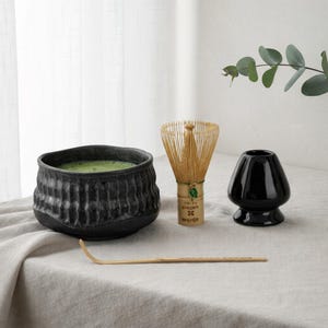 May include: A matcha tea set featuring a black ceramic bowl with green tea, a bamboo whisk, a black ceramic whisk holder, and a bamboo scoop. The set is arranged on a neutral-colored linen surface, with a sprig of greenery in the background.