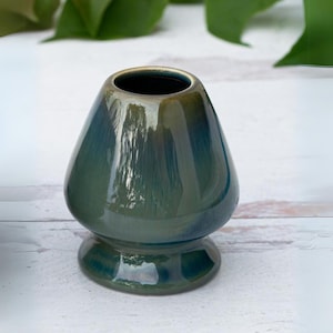 May include: A teal-colored ceramic matcha tea bowl with a glossy finish. The bowl has a rounded body and a flared rim with a gold accent. The base is a circular pedestal. The bowl is designed for whisking matcha tea.