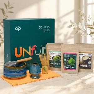 May include: A matcha tea set including a dark green box labeled UNfurl, a blue ceramic bowl, a blue ceramic whisk holder, a bamboo whisk, a metal strainer, and three pouches of organic matcha and hojicha powder by Oprka on an orange cloth.