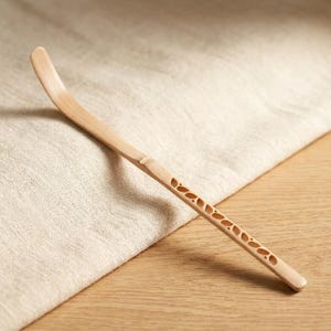 May include: A light-colored bamboo tea whisk, also known as a chasen, with a curved handle and a section of decorative cut-out leaf shapes. The whisk rests on a light-colored cloth and wooden surface.