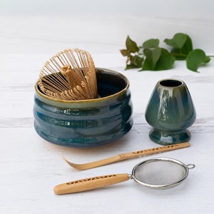 Puede incluir: Un juego de té matcha que incluye un cuenco de cerámica azul profundo, un soporte para batidor azul a juego, un batidor de bambú, una cuchara de bambú con patrón recortado y un colador de metal con mango de madera con patrón recortado.