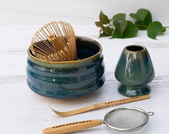 以下が含まれることがあります： 深い青色の陶器製ボウル、お揃いの青い茶筅立て、竹製の茶筅、切り抜き模様のある竹製の茶杓、そして同じ切り抜き模様が施された木製ハンドルの金属製茶こしを含む抹茶セット。