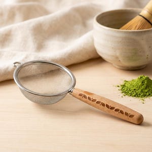 May include: A stainless steel tea strainer with a wooden handle featuring a leaf design. The strainer is next to a bowl, a whisk, and a pile of green matcha powder, suggesting a tea preparation theme.
