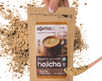 Puede incluir: Una bolsa de papel marrón con una etiqueta blanca que dice "aprika" y "hojicha" en texto negro. La bolsa contiene polvo de té verde tostado orgánico. La etiqueta también incluye el texto "Yabukita Cultivar" y "Hecho en Japón".