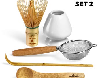 May include: A matcha tea set with a bamboo whisk, a white ceramic bowl, a stainless steel strainer, a bamboo scoop, and a wooden spoon. The set includes the text "SET 2". The whisk has a green band and a gold base with text.