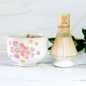 May include: A white ceramic tea bowl with a floral pattern in pink and yellow.  A bamboo matcha whisk with a white base and a green string is next to the bowl. The text "oprea  by/tea  pajun" is printed on the whisk handle.