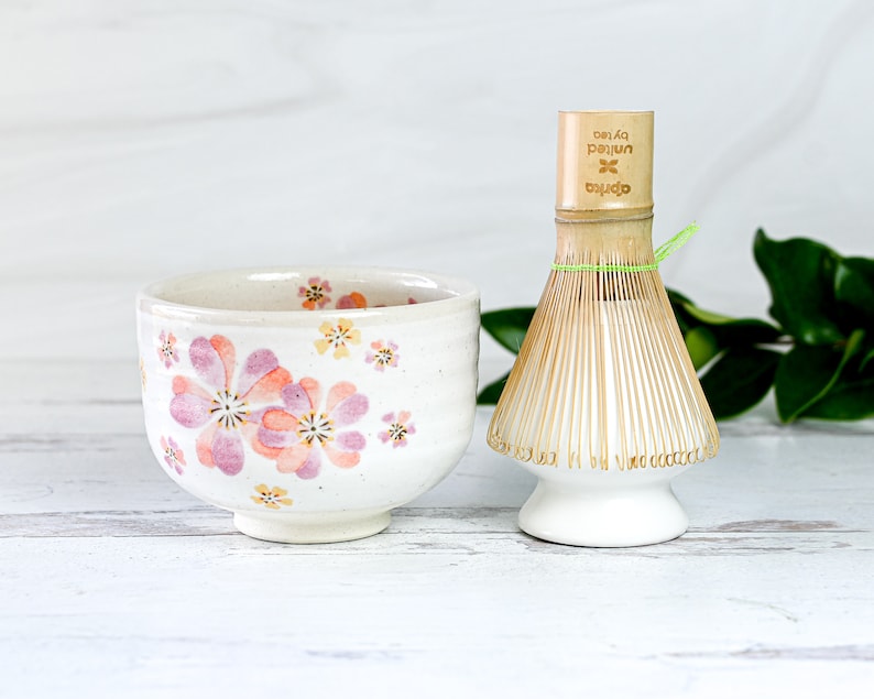 May include: A white ceramic tea bowl with a floral pattern in pink and yellow.  A bamboo matcha whisk with a white base and a green string is next to the bowl. The text "oprea  by/tea  pajun" is printed on the whisk handle.