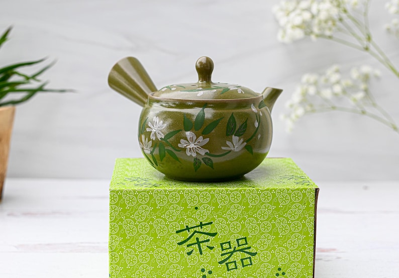 Puede incluir: Una tetera de cer&aacute;mica verde con un dise&ntilde;o floral blanco. La tetera est&aacute; sentada sobre una caja verde con un patr&oacute;n blanco y el texto "茶器" en negro.