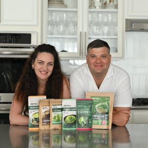 Puede incluir: Una pareja posa con varios paquetes de productos de t&eacute; matcha. Los paquetes var&iacute;an en tama&ntilde;o y dise&ntilde;o, con tonos verdes y marrones. La marca "aprika" es visible en el embalaje. El fondo es una cocina moderna con gabinetes blancos y electrodom&eacute;sticos de acero inoxidable.
