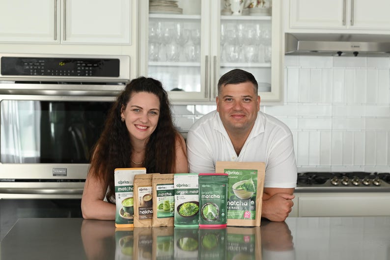Puede incluir: Una pareja posa con varios paquetes de productos de t&eacute; matcha. Los paquetes var&iacute;an en tama&ntilde;o y dise&ntilde;o, con tonos verdes y marrones. La marca "aprika" es visible en el embalaje. El fondo es una cocina moderna con gabinetes blancos y electrodom&eacute;sticos de acero inoxidable.