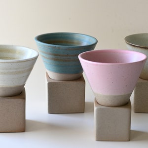 May include: Four ceramic bowls in varying colors: cream, teal, pink, and beige. Each bowl has a light-colored base and sits atop a square, neutral-toned pedestal. The bowls have a conical shape.